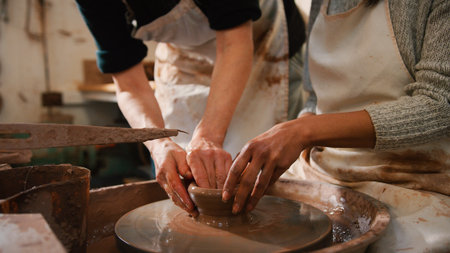 Close Up Of Male Teacher Helping Woman Sitting At Wheel In Pottery Classの写真素材
