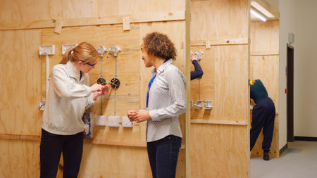 Female Tutor With Trainee Electricians In Workshop Studying For Apprenticeship At College