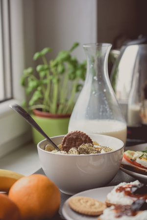 White bowl of cornflakes, slice of banana and chocolate on top. Breakfast set in background with milkの写真素材