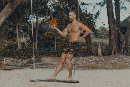 Young handsome man sitting on the beach with pandan fruit in hand. Shirtless wearing black boxer...
