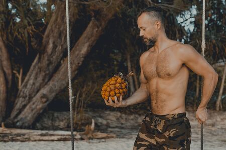 Young handsome man sitting on the beach with pandan fruit in hand. Shirtless wearing black boxer...