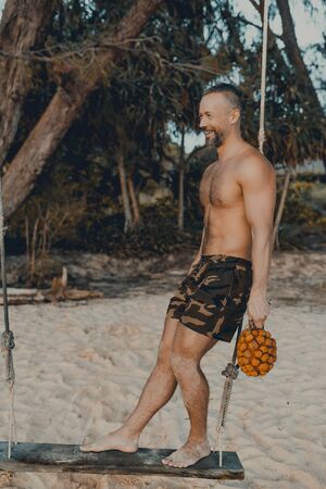 Young handsome man sitting on the beach with pandan fruit in hand. Shirtless wearing black boxer shorts, showing muscular fit body. Koh Rong Samloem island, Lazy beach. Cambodia, Asiaの写真素材