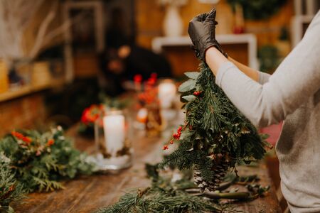 Christmas workshop. Hands holding fir branches, pine cones, berries, thread, scissors on wooden table. Winter holidays celebration conceptの写真素材