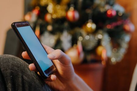 Hand holding smartphone in front of decorated christmas tree, christmas concept. seasonal greetingsの写真素材
