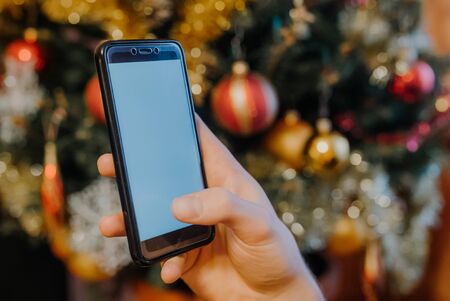 Hand holding smartphone in front of decorated christmas tree, christmas concept. seasonal greetings