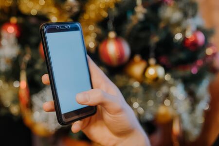 Hand holding smartphone in front of decorated christmas tree, christmas concept. seasonal greetingsの写真素材