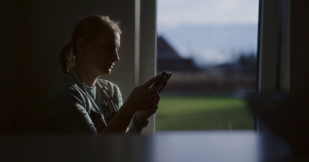Silhouette of a woman browsing smartphone in front of a kitchen windowの写真素材