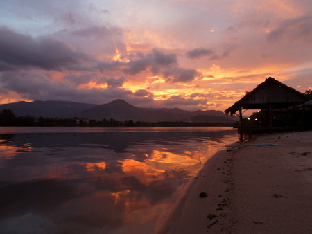 Sunset along a jungle river in Cambodia, with grass and wood hut visible in foreground and distant mountains.の写真素材