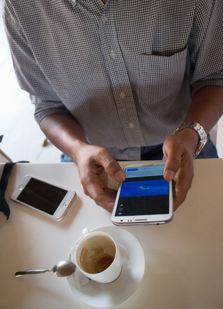 High angle medium shot of a male person of color looking at a smartphone 'Phablet' in a cafe-like environment. A coffee cup and another smartphone can be seen nearby.の写真素材