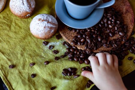 child's hand reaches for coffee beans scattered on the table, a cup with coffee and muffins nextの写真素材