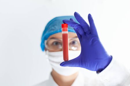 female doctor in safety glasses, mask and gloves looks at a test tube with bloodの写真素材