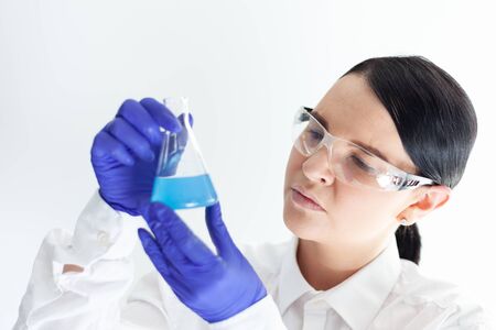 female scientist with protective eyeglasses and gloves holding a flask with blue liquid substanceの写真素材