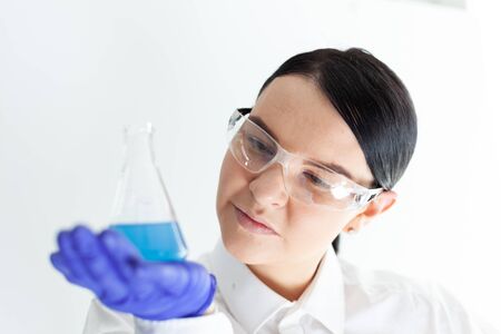 Female scientist with protective eyeglasses and gloves holding a flask with blue liquid substance in...