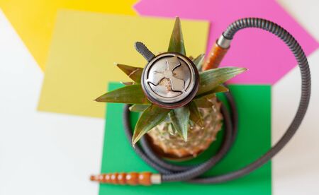 Hookah made from ripe pineapple fruit on a white table, shisha, yellow pink green background