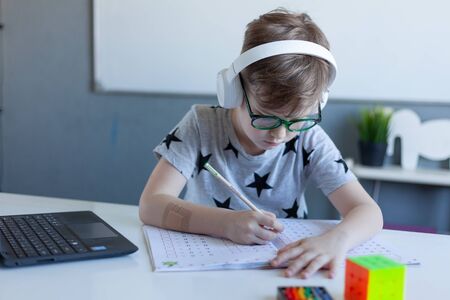 Boy is engaged at home online school, learning in isolationA boy writing down in a notebook was taken out of a laptop. The child is remotely engaged using a laptop. Distance Learning during Quarantine Conceptの写真素材