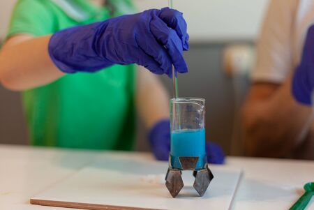 close-up of a child's hand in medical gloves with a glass rod stirs blue liquid in a glass flask