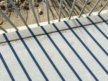sunlight through a while metal fence with shadows casting on concrete sidewalk diagonal view