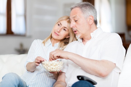 Couple watching tv and eating popcorn on the sofa at home in living roomの写真素材