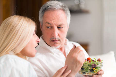 Mature couple talking and eating a salad while sitting in their living roomの写真素材