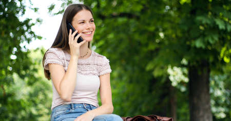 Portrait of a happy woman sitting on the bench and talking on the phone outdoorsの写真素材