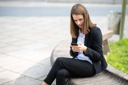 Business woman using her mobile phone while sitting on a bench in front of her officeの写真素材