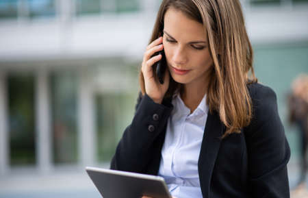Portrait of a young woman using a tablet while talking on her mobile cell phone in front of her officeの写真素材