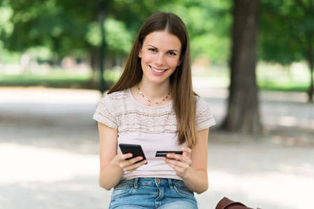 Woman doing online shopping using her credit card while sitting in a parkの写真素材