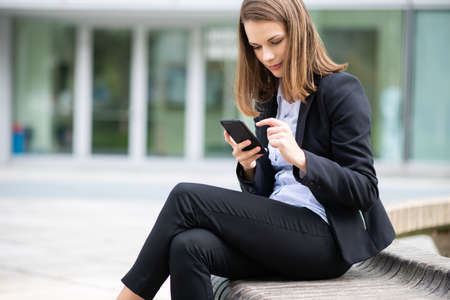 Business woman using her mobile phone while sitting on a bench in front of her officeの写真素材