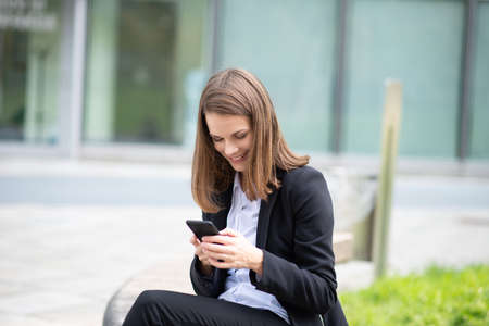 Business woman using her mobile phone while sitting on a bench in front of her officeの写真素材
