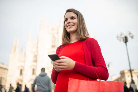 Smiling shopping woman walking in a city while using her smartphoneの写真素材