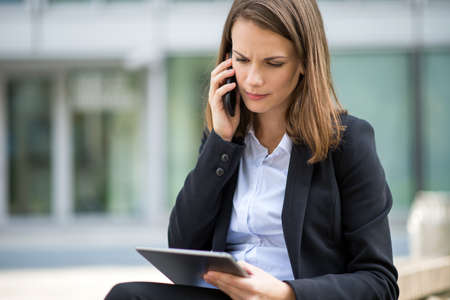 Portrait of a young woman using a tablet while talking on her mobile cell phone in front of her officeの写真素材