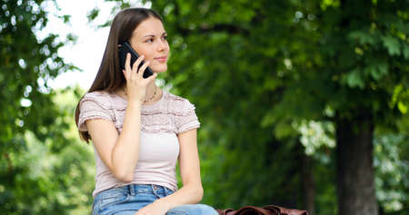 Portrait of a happy woman sitting on the bench and talking on the phone outdoorsの写真素材