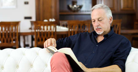 Mature man relaxing while reading a book on a sofa at homeの写真素材