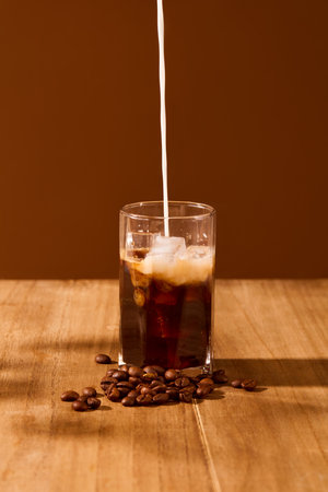 A stream of milk is pouring from above into a cup of black coffee on a wooden table with a brown...