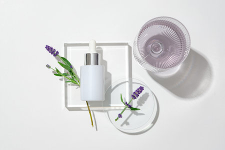 View from above of a white serum bottle on a glass podium, lavender flowers on a petri dish and a glass of water on a white backdrop. Blank labels for branding.の写真素材