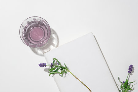 A glass containing a transparent light purple liquid, a white podium and fresh lavender flowers displayed on a minimalist white background. Space for advertising.の写真素材