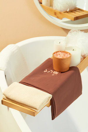 Close-up of a wooden tray placed across the bathtub with cloth towels, pink salt, scented candles and a bath sponge. Space with two main colors white and brown. Relax inside the bathroom.の写真素材