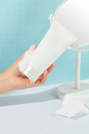 A hand holding a tube of unbranded facial cleanser with white foam, a mirror and cotton pads on a...