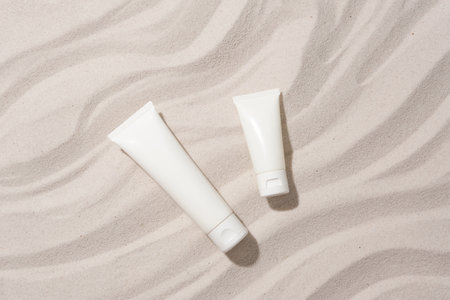 Close-up of two unlabeled cosmetic tubes placed on a sandy background with wave patterns. Mockup for...