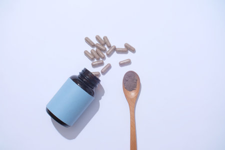 Silver hard capsules are poured onto the table from an unbranded pill bottle, next to it is a wooden spoon displaying a handful of powder against a pastel blue background. Scene for drug advertising.の写真素材