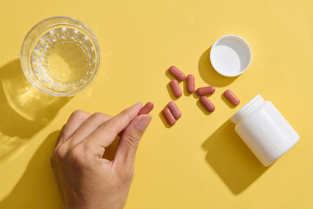 A hand holding pill, a glass of water, and an unlabeled medicine bottle on a yellow background. Medication and prescription pills concept.の写真素材
