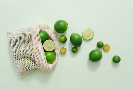A mesh bag containing a lot of fresh lime and kumquat displayed on light background. Lime and...
