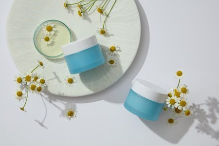 Flat lay of two unlabeled blue jars arranged with glass petri dish and flowers. Chamomilla (Matricaria chamomilla) is very useful in cosmetic productionの写真素材