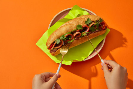 Two woman hands holding a golden knife and fork preparing to eat bread with filling wrapped green paper on round pink plate on orange background. Street food of Vietnamese, advertising photoの写真素材