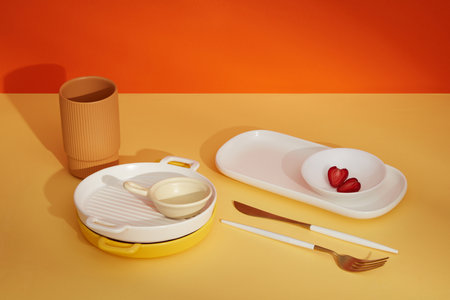 Stacked of pan, white plates, cutlery and strawberry slices decorate on color background. Modern...