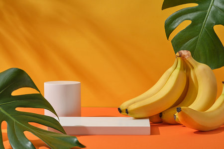 Fresh bunch of banana, jungle monstera leaves and cylinder white podium decorated on a yellow background with natural shadow leaves. Space for display cosmetic of banana extractの写真素材