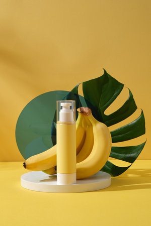 An empty pump bottle placed on round podium with fresh bananas and green leaf on yellow background. Mockup for design, organic cosmetics, beauty products conceptの写真素材