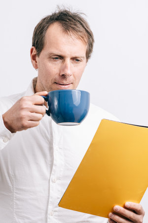 Young man looking at a yellow folder drinking coffee in a blues cup. White background.