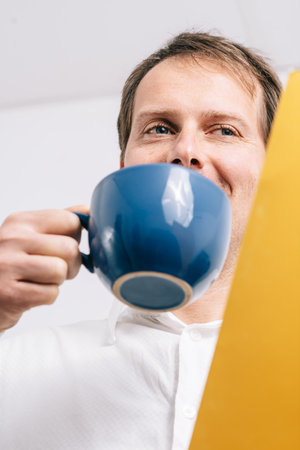 Young man looking at a yellow folder drinking coffee in a blues cup. White background.