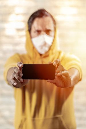 Close up of man with mask showing smartphone wearing yellow hooded .. Shining light.の写真素材